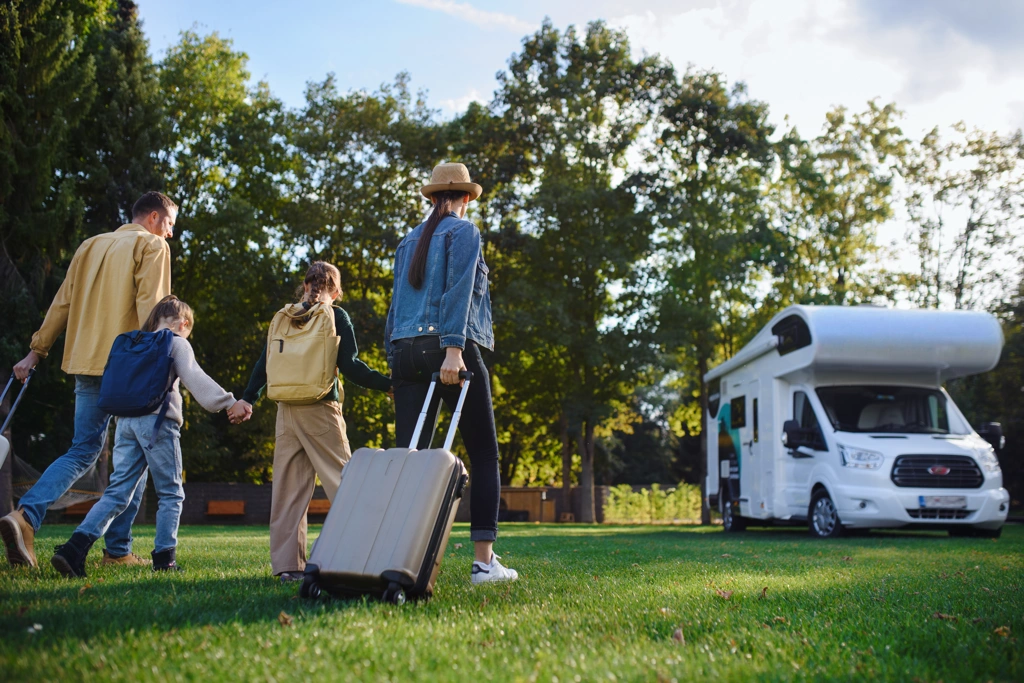 Famille marchant dans un parc verdoyant avec des valises en direction d’une roulotte stationnée, illustrant l’esprit de liberté et les idées de voyages en VR pour 2026.