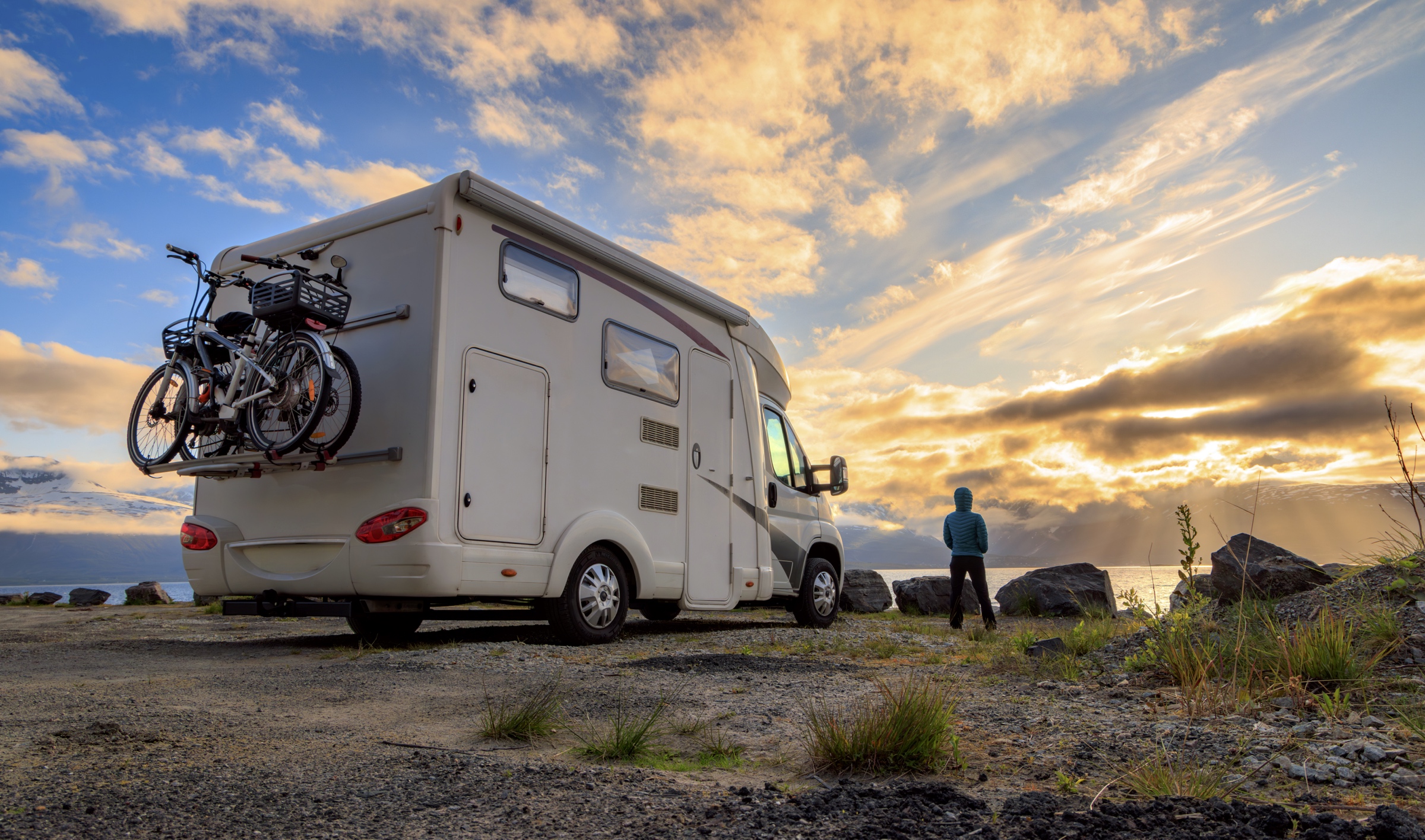 Motorisé classe C stationné près d’un lac au coucher du soleil, avec deux vélos à l’arrière et une personne observant le paysage.