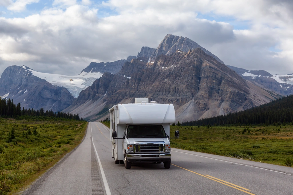 VR blanc roulant sur une route des Rocheuses bordée de montagnes majestueuses, représentant l’aventure et la liberté d’un road trip à travers le Canada.