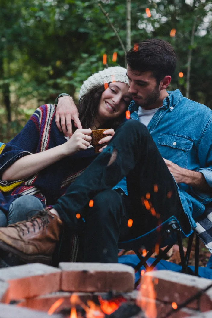Couple assis près d’un feu de camp dans un décor forestier, partageant une boisson chaude lors d’un séjour en roulotte, illustrant des vacances romantiques en plein air pour deux.