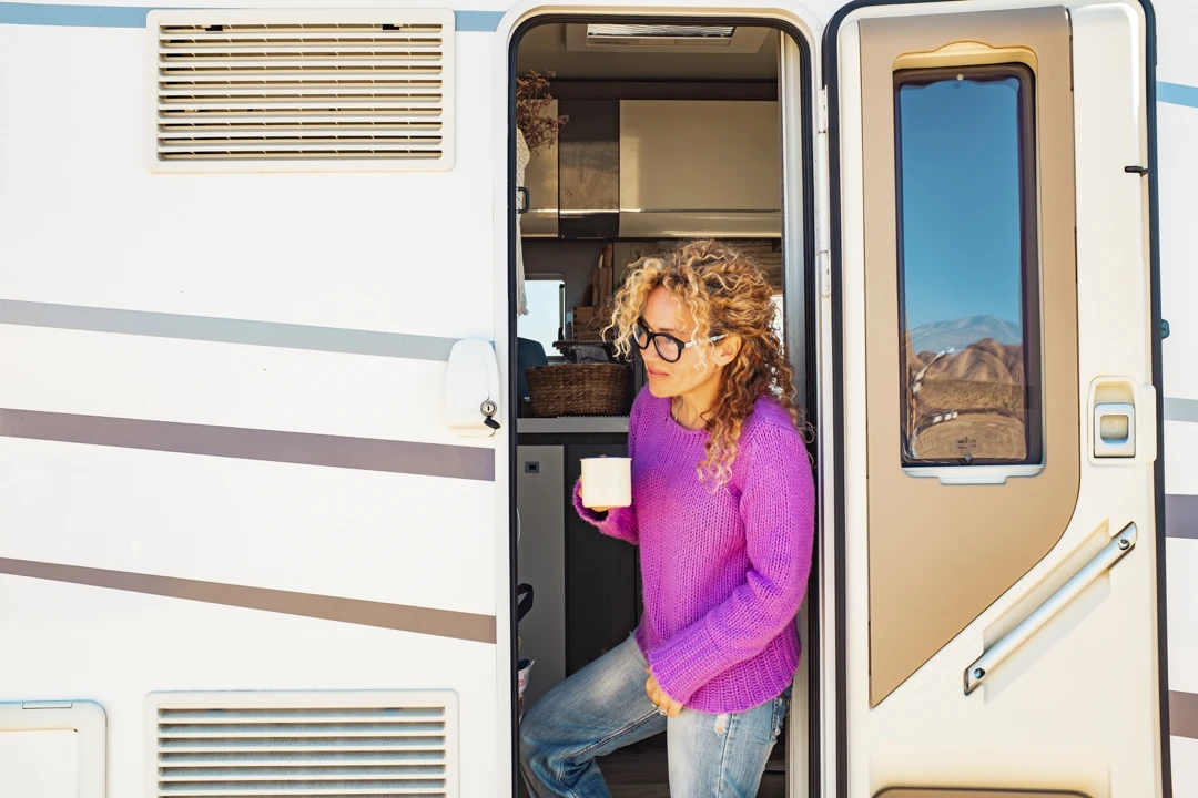 Femme en chandail mauve sortant de son VR avec une tasse à la main, profitant du paysage gaspésien en arrière-plan.