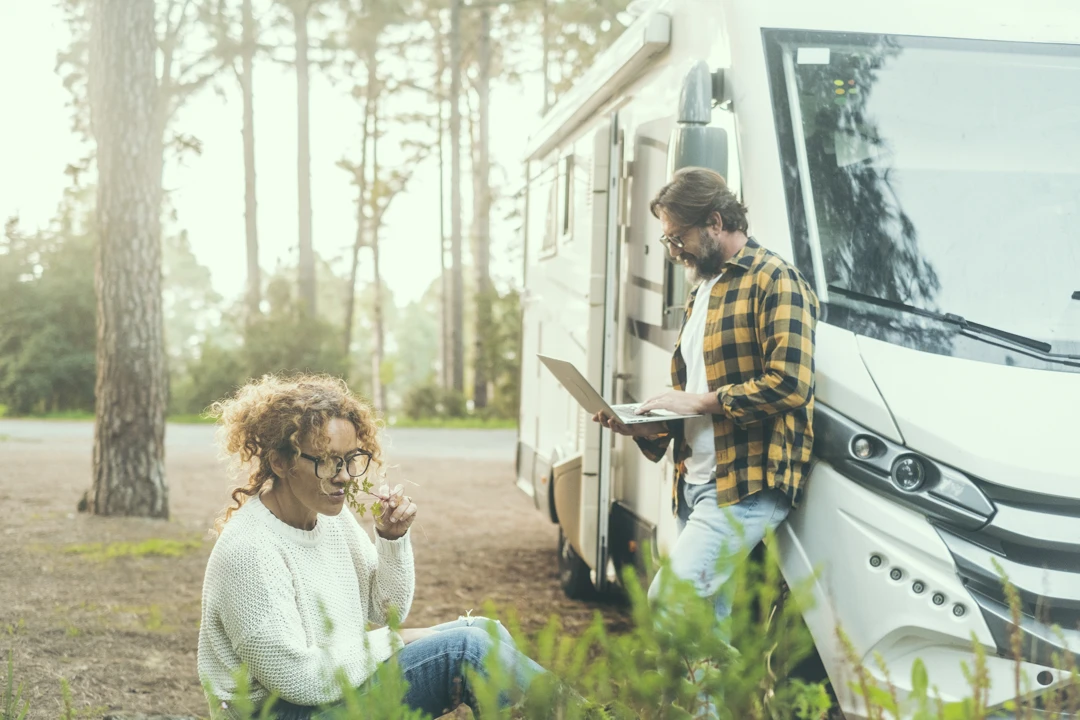 Scène matinale dans un camping en Gaspésie : femme assise au sol parmi les plantes, homme debout avec un ordinateur portable près d’un véhicule récréatif.