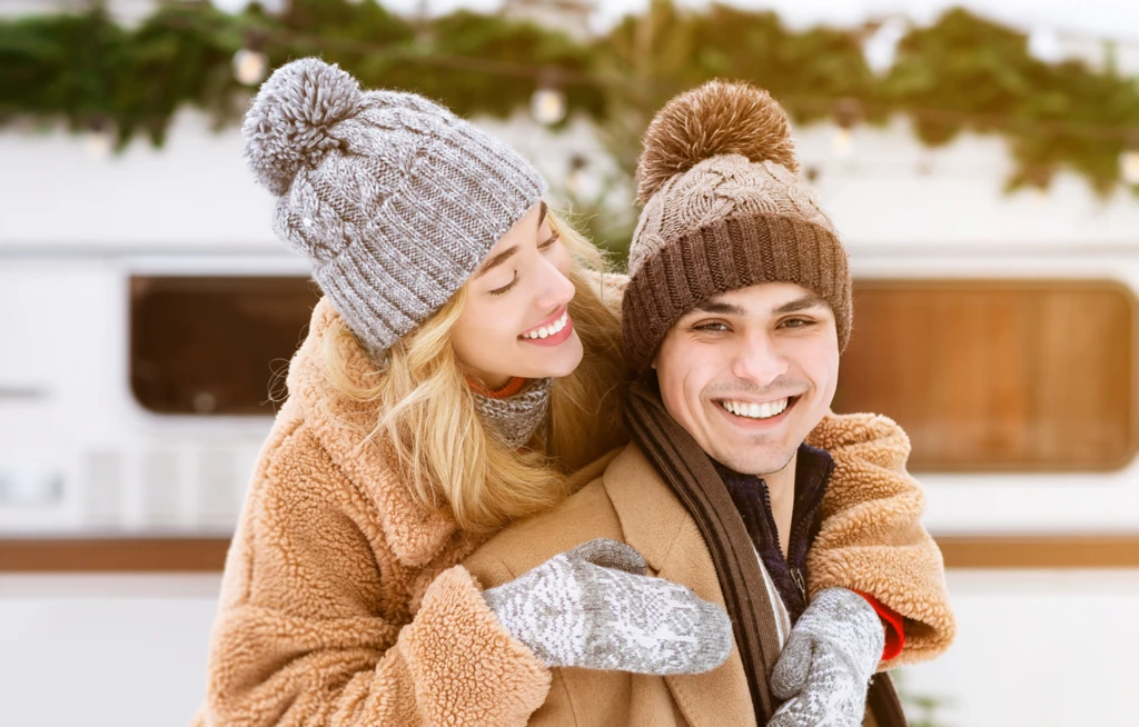 Couple souriant habillé pour l’hiver devant une roulotte décorée, illustrant les avantages d’acheter une roulotte en hiver pour planifier la prochaine saison de voyage.