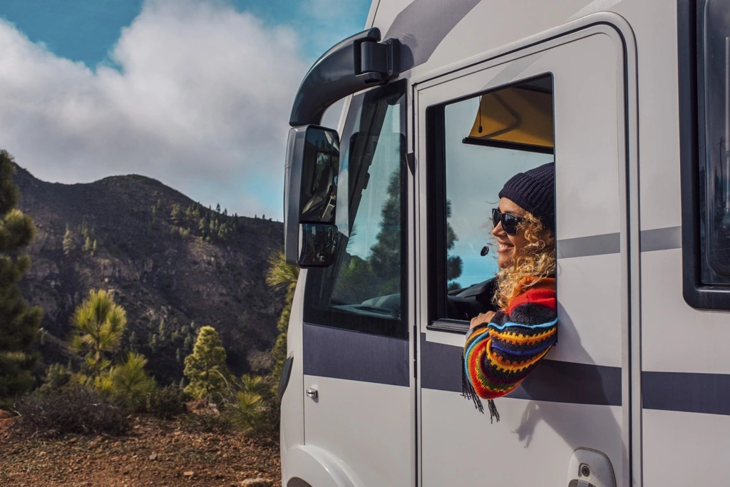 Femme souriante appuyée à la fenêtre d’un VR stationné en montagne, représentant la liberté et l’esprit d’aventure d’un road trip à travers le Canada.