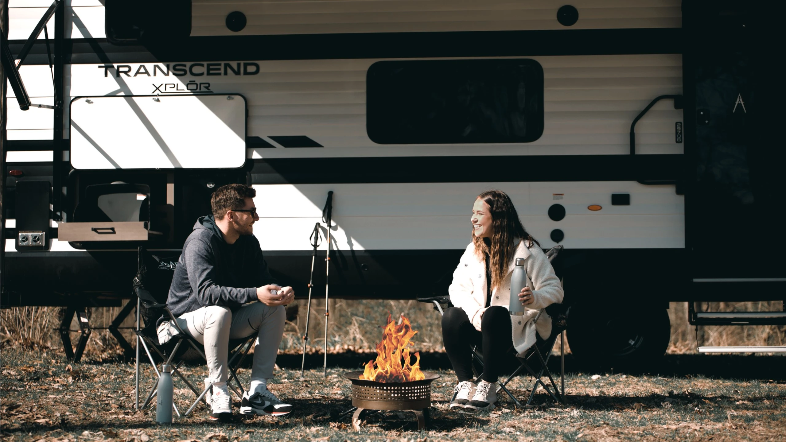 Couple assis devant un feu de camp près d’une roulotte Transcend Xplor, illustrant la durabilité et la résistance aux infiltrations des modèles Quadseal de Grand Design.